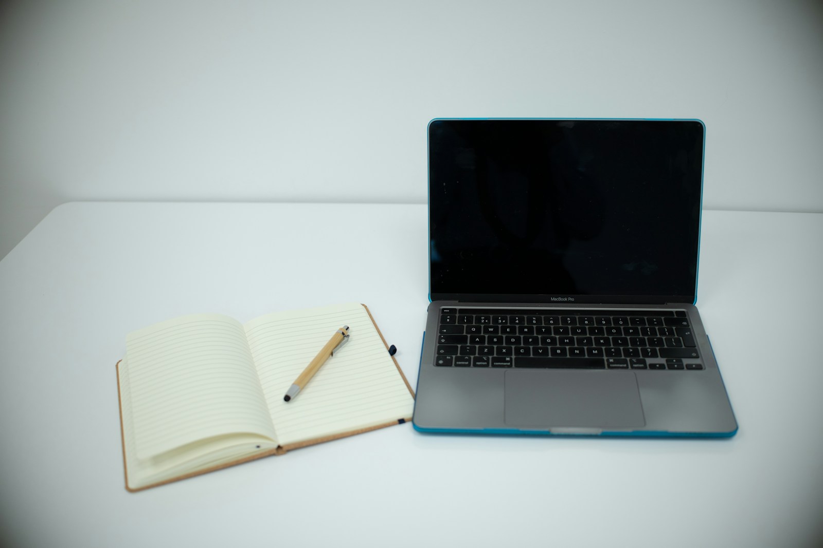 Een laptop naast een open notitieboek en een pen op een bureau.